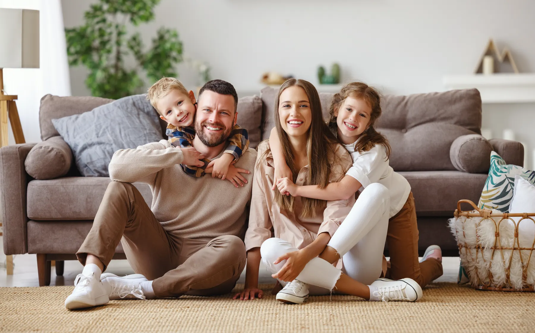 A smiling family of four sitting on the rug in front of their sofa